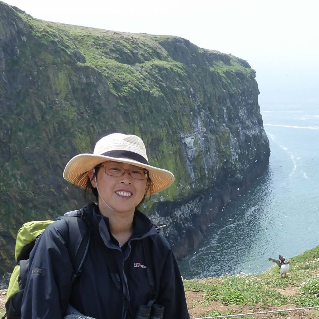 Head and shoulder shot of me on Skomer Island.  Wearing sunhat, raincoat and rucksack near seacliffs, with sea and inlet behind.  Puffin standing behind me with wings flapping.